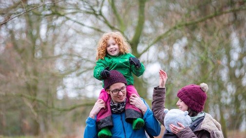 Family of four - toddler on her father's shoulders, and mother with a baby in a papoose, walking through the woodland at Acorn Bank.